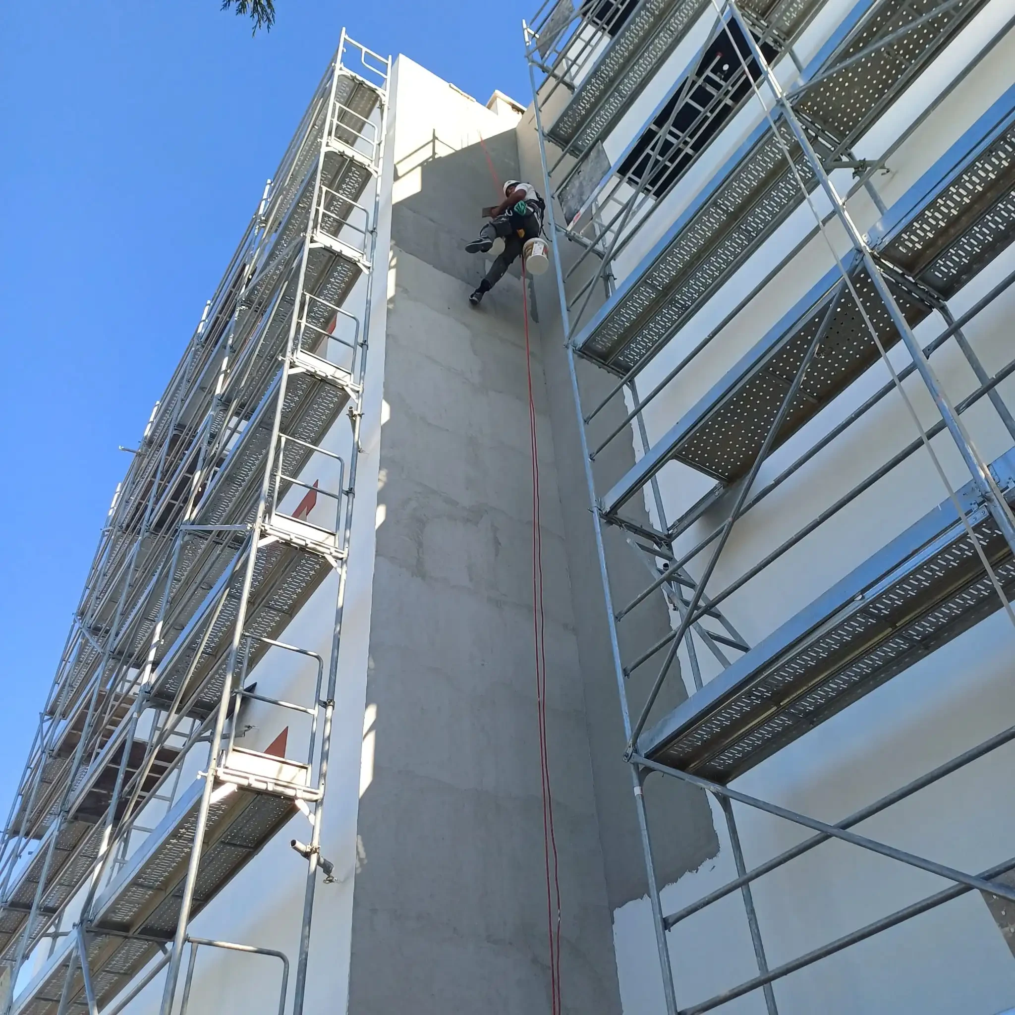 Técnico em trabalhos verticais, suspenso por cordas, a reparar a fachada de um prédio entre andaimes.